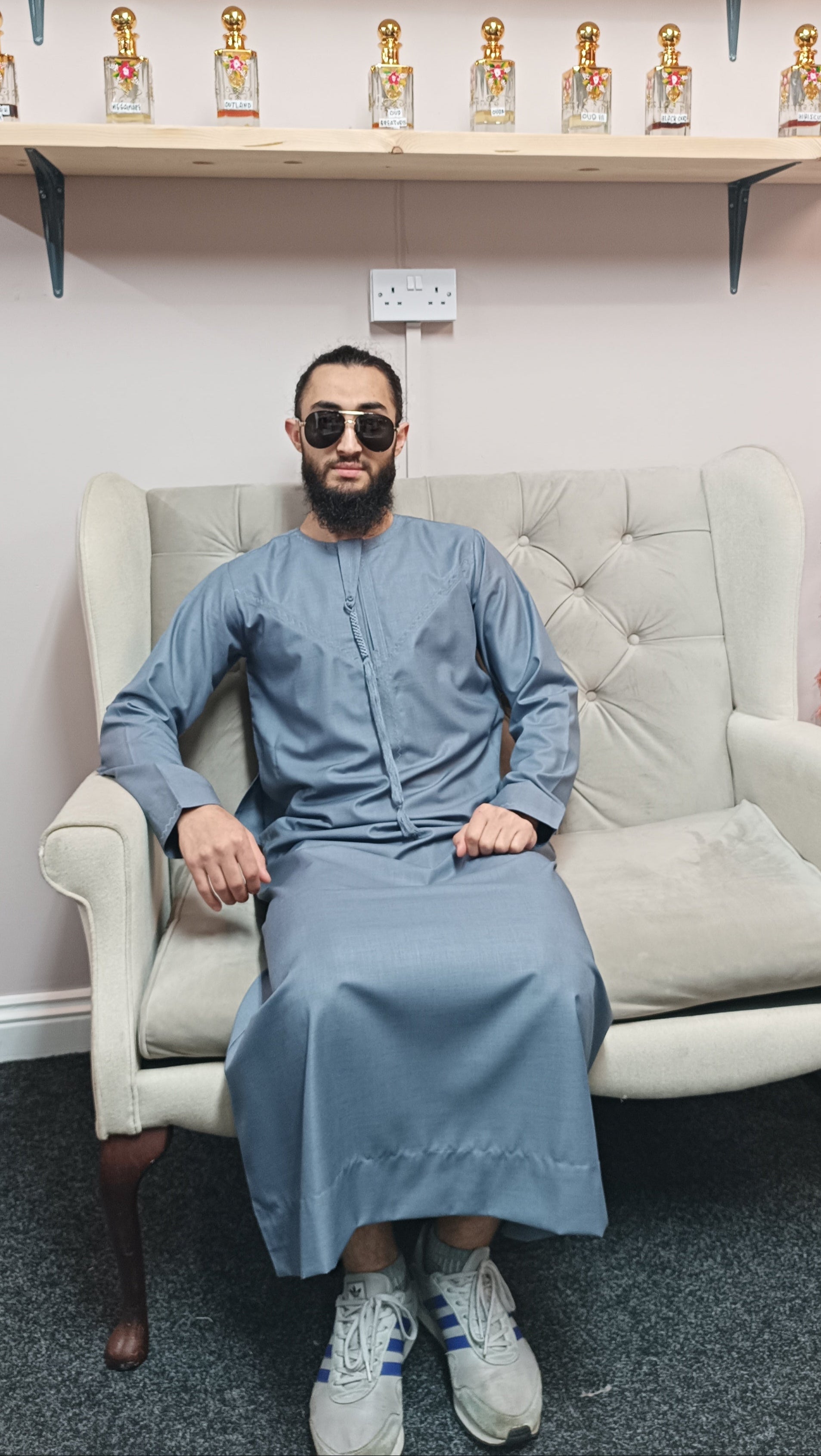 Man sitting on a white chair wearing sunglasses and a light blue outfit, with shelves of perfume bottles in the background.